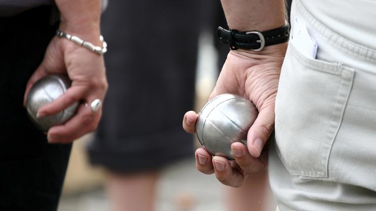 Zwei Personen halten silberne Kugeln in der Hand, während sie auf einem Platz an einem Boule-Spiel teilnehmen.