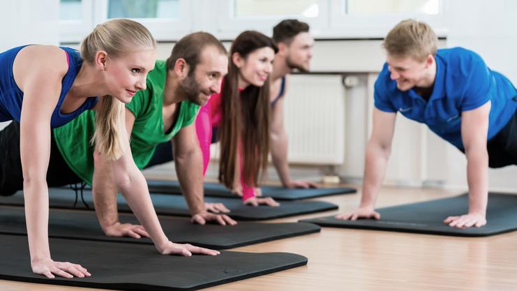 Fünf Personen in Sportbekleidung machen Liegestütze auf Matten in einem hellen Fitnessraum.