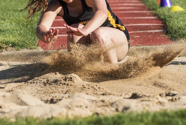 Athletin springt in einen Sandgruben, während Sand aufwirbelt. Grüße des Sprungbretts sind im Hintergrund sichtbar.