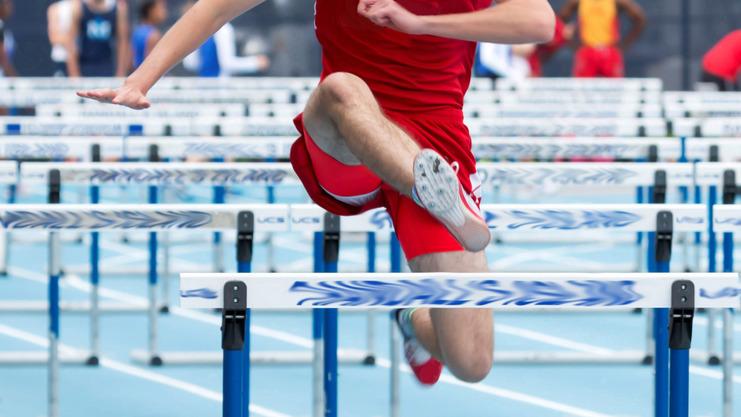 Ein Athlet springt über eine Hürde auf einer blauen Leichtathletikbahn während eines Wettkampfs.
