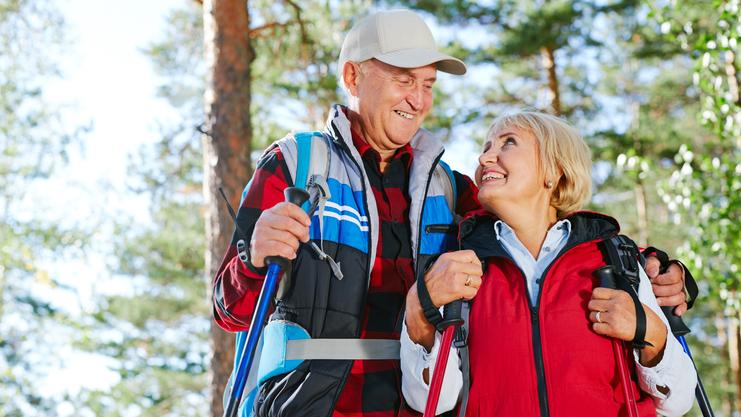 Ein älteres Paar steht in einem Wald, lächelt sich an und hält Wanderstöcke in den Händen.