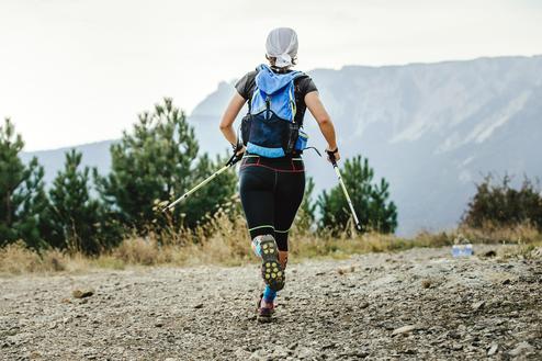 Frau mit Wanderstöcken läuft auf einem Schotterweg, im Hintergrund sind Berge und Bäume sichtbar.