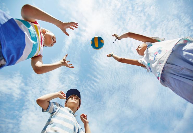 Drei Kinder werfen einen bunten Volleyball in den Himmel vor einem blauen Himmel mit einigen Wolken.